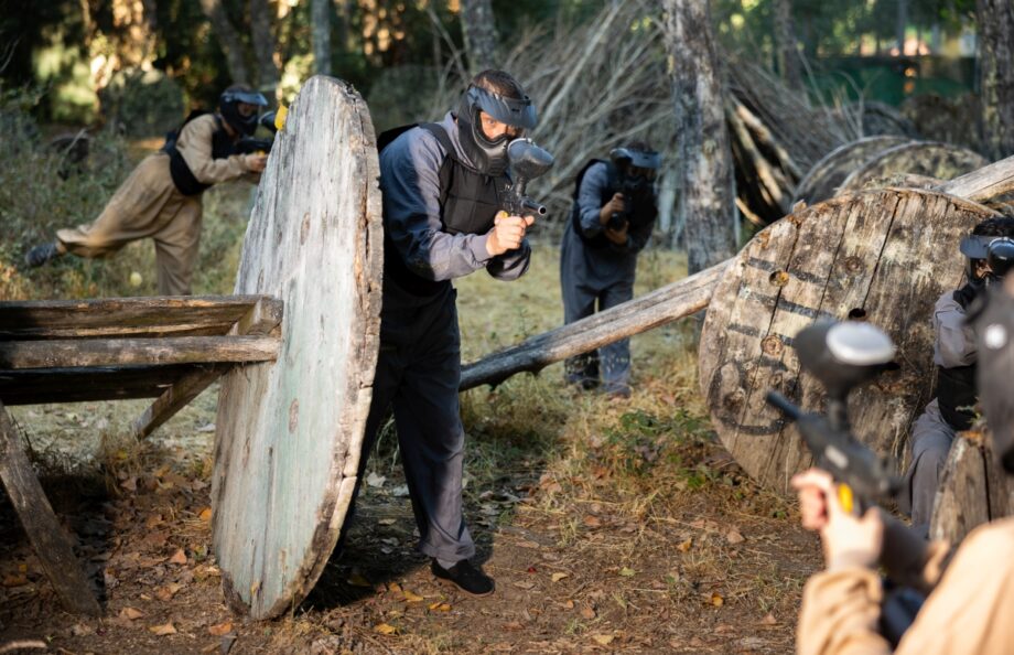 Grupa osób uczestniczy w grze na pole paintballowe, kryjąc się za przeszkodami w lesie.
