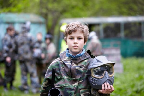 Młody chłopiec w stroju maskującym trzyma kask podczas wydarzenia "Paintball dla szkół".