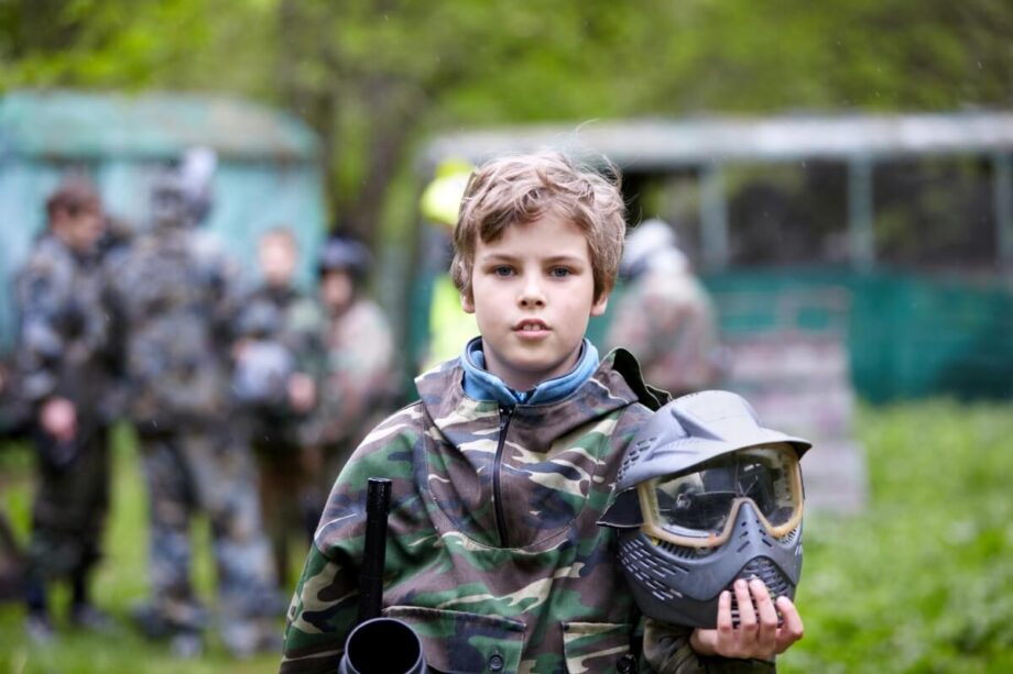 Młody chłopiec w stroju maskującym trzyma kask podczas wydarzenia "Paintball dla szkół".