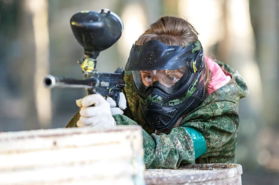 Kobieta w masce ochronnej celuje z pistoletu, biorąc udział w paintballu na wieczór panieński.